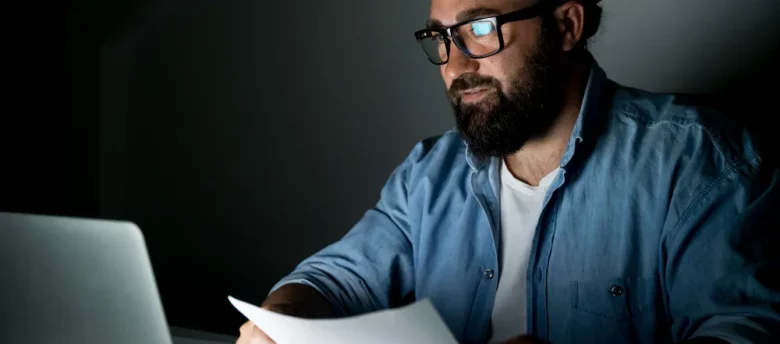 Homem em quarto escuro com papéis na mão checando notebook - consultar nome sujo