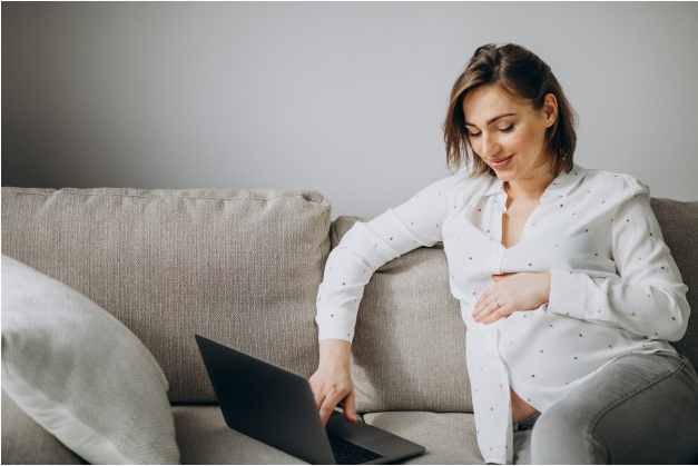 mulher grávida usando camisa branca deitada ao sofá usando um laptop enquanto acaricia sua barriga