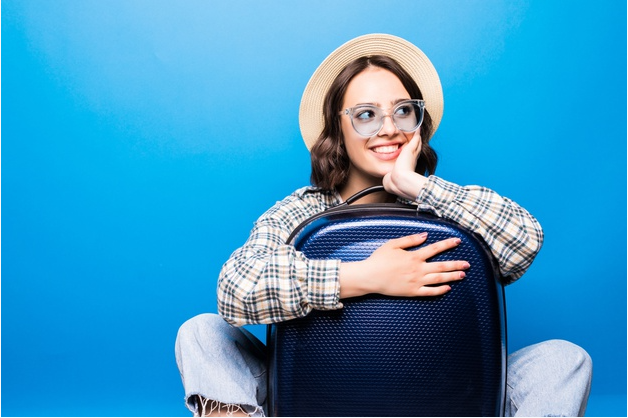 mulher usando chapéu panamá branco abraçando mala de viagem azul e sorrindo de costas para uma parede azul