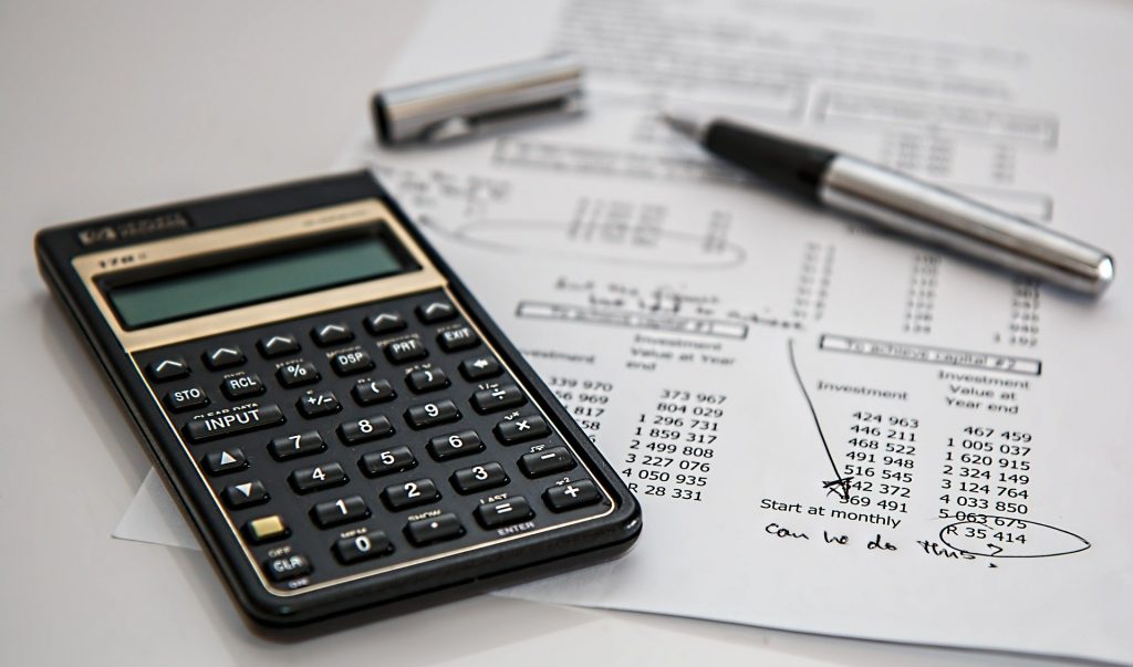 Calculadora, papel com dados monetários e caneta em uma mesa branca.