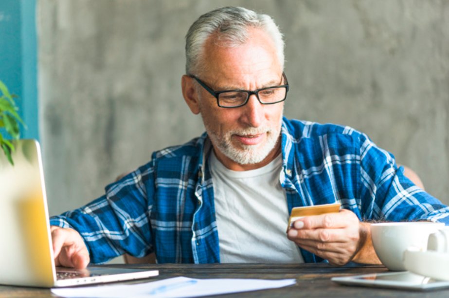 senhor de idade usando camisa azul mexendo em seu laptop e segurando um cartão de crédito 
