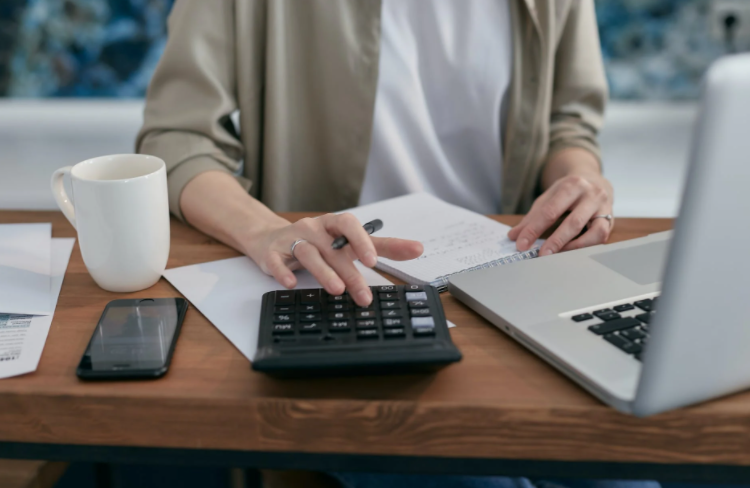 mulher sentada à mesa mexendo na calculadora com bloco de notas, caneca e computador à frente 