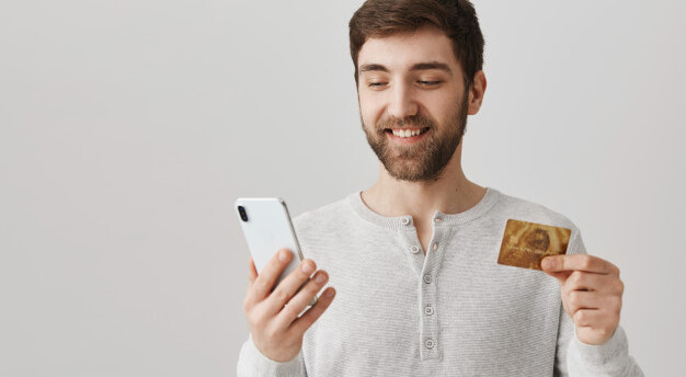 homem de camiseta branca sorri enquanto segura um celular e um cartão de crédito