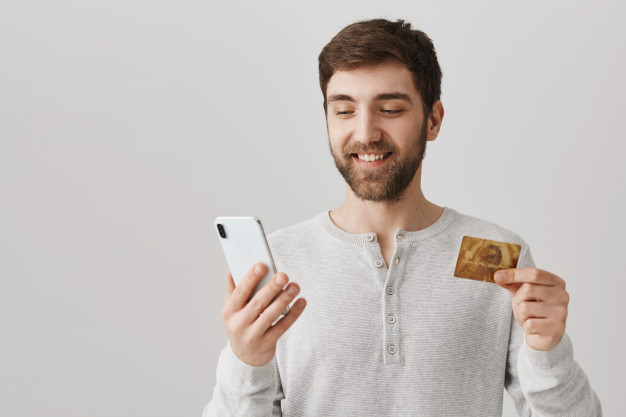 homem de camiseta branca sorri enquanto segura um celular e um cartão de crédito