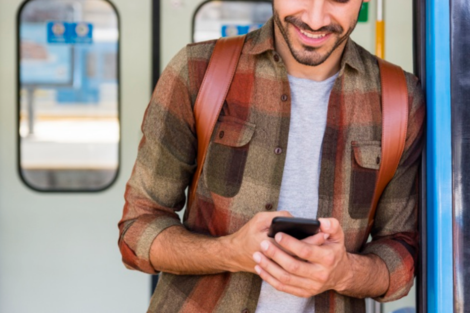 homem de camisa xadrez marrom apoiado em trem com uma mochila nas costas e mexendo em seu celular