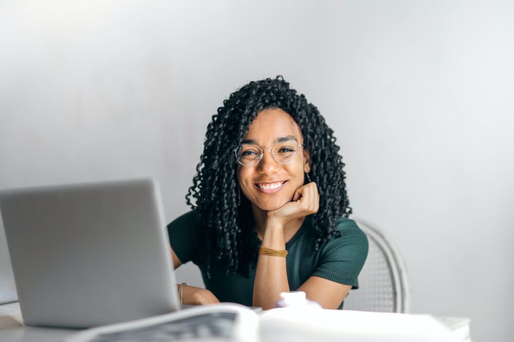 mulher de óculos e camiseta verde sentada a uma mesa de frente para um laptop sorri com a cabeça apoiada na mão