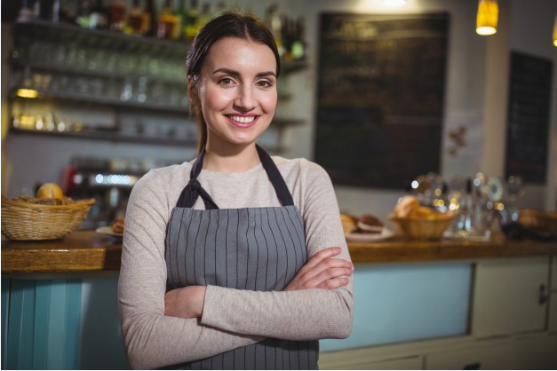 imagem de mulher usaando camisa e manga comprida e avental cruzando os braços e sorrindo dentro de seu restaurante
