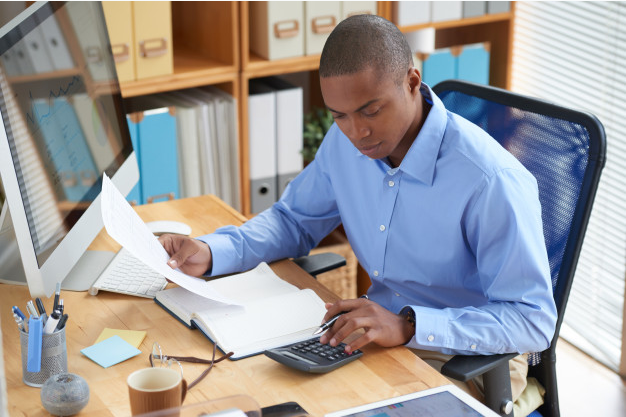 homem de camisa azul sentado a uma mesa com um computador usando uma calculadora e segurando uma folha de papel