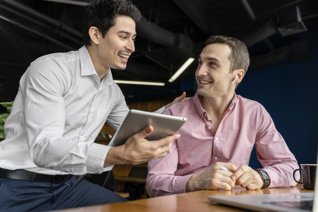 dois homens de camisa conversam sendo um sentdo a uma mesa e o outro de pé com um tablet em mãos
