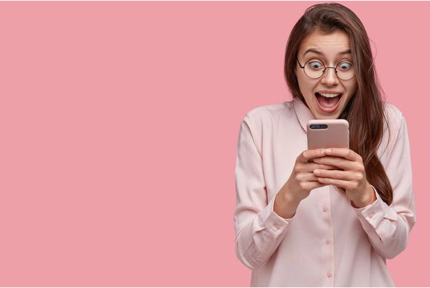 mulher de camisa e óculos sorri enquanto olha para seu celular de costas para um fundo rosa