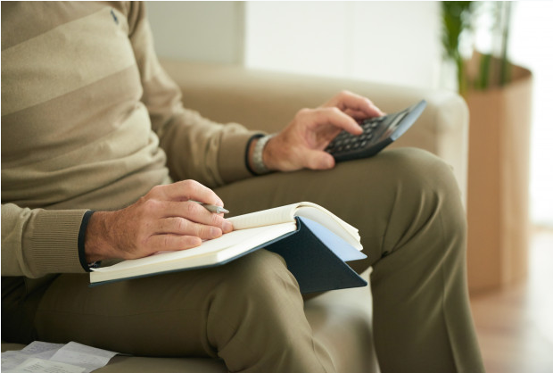 imagem ampliada de idoso sentado em poltrona fazendo contas com uma calculadora apoiada em sua perna esquerda e anotações em um caderno que está em sua perna direita