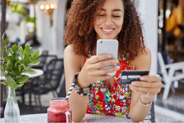 jovem sentada a uma mesa em um estabelecimento sorri enquanto usa seu celular e segura um cartão de crédito