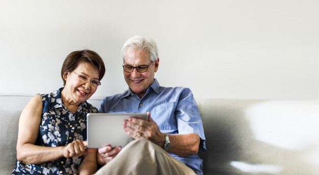 casal de idosos sentados em sofá branco sorriem enquanto usam um tablet