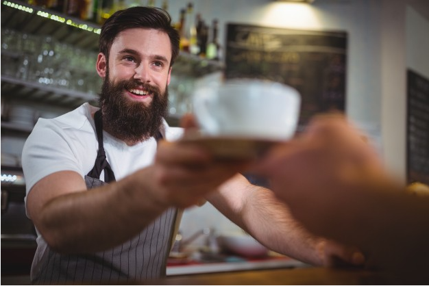 homem com barba usando camiseta branca e avental escuro sorri enquanto serve uma xícara branca de café à outra pessoa