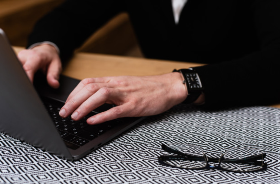 homem de roupas pretas mexendo em seu laptop sentado a uma mesa de madeira com seu óculos de garu na lateral