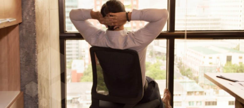 empresário de roupa social sentado em sua cadeira dentro de sua sala com as braços cruzados atrás da cabeça e reclinando para trás
