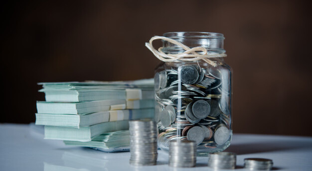 imagem de jarro com moedas, maços de notas em cima de uma mesa branca