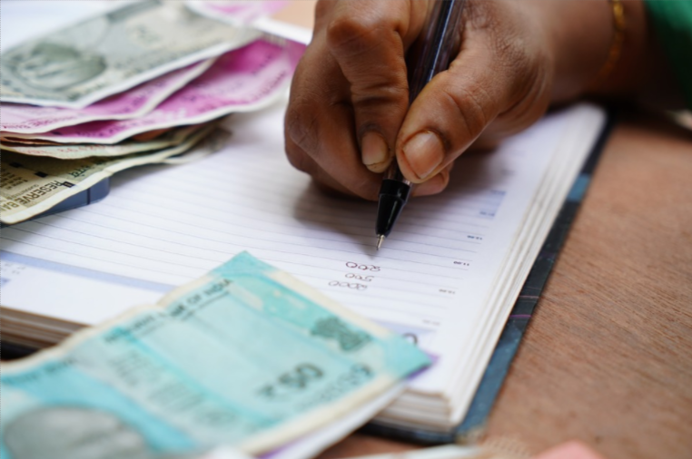Mão de uma pessoa segurando uma caneta e fazendo anotações sobre como pagar dívidas acumuladas em uma agenda rodeada de notas de dinheiro.