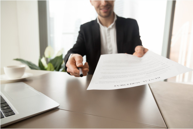 homem de terno e camisa branca sentado a uma mesa de madeira com um laptop oferece um contrato e uma caneta para assinar