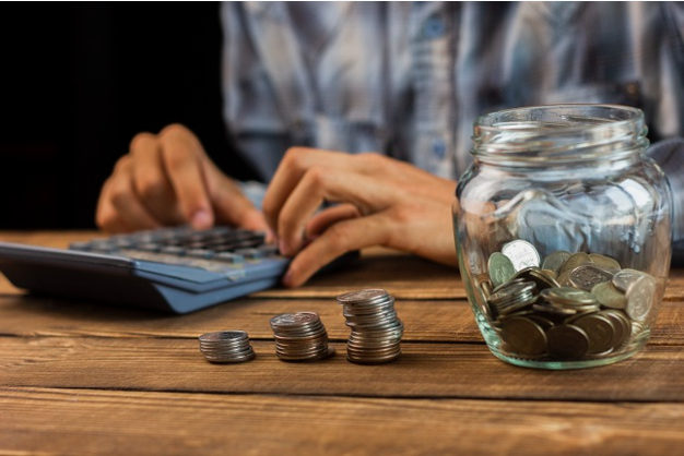 homem sentado a uma mesa usando uma calculadora em frente uma pote com moedas ao lado de pilhas de moedas
