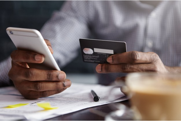 imagem amppliada de homem de camisa branca segurando um cartão de crédito com uma mão e usando um celular com outra enquanto está sentado a uma mesa com contas a pagar