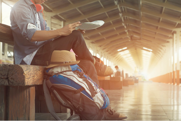 homem com mapa e bagagem de mochileiro aguarda sentado um trem em uma plataforma de embarque
