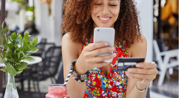 imagem de mulher apoiada em uma mesa sorrindo e segurando um cartão de crédito e um celular em suas mãos