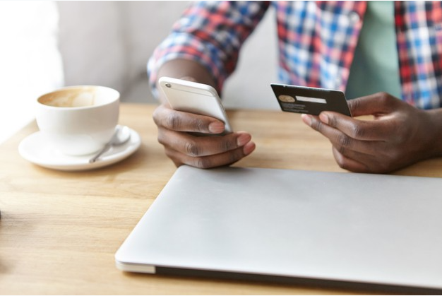 imagem ampliada de um homem de camisa xadrez sentado a uma mesa com uma xícara de café e um laptop enquanto segura um cartão de crédito e um celular