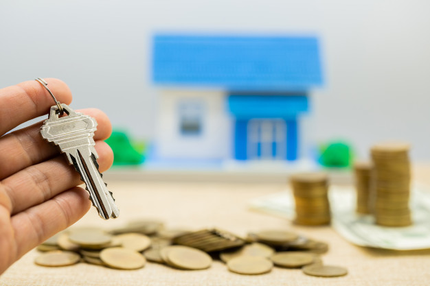 mão segurando molho de chaves à frente de montes de moeda e uma casa em miniatura em cima de uma mesa