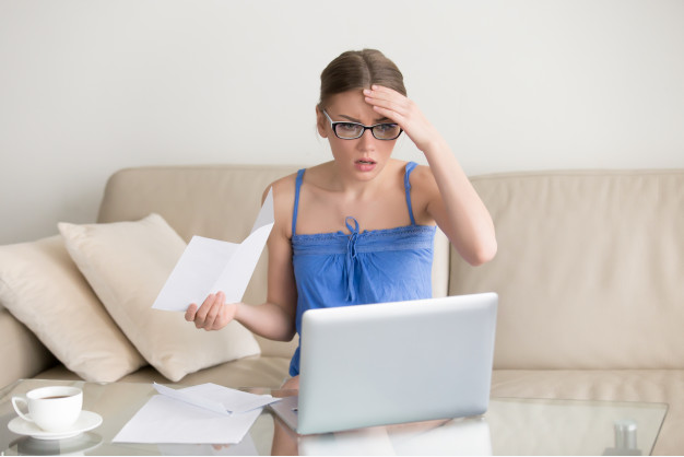 mulher loira de óculos e camisa azul sentada em um sofá de frente para u computador com semblante preocupado 