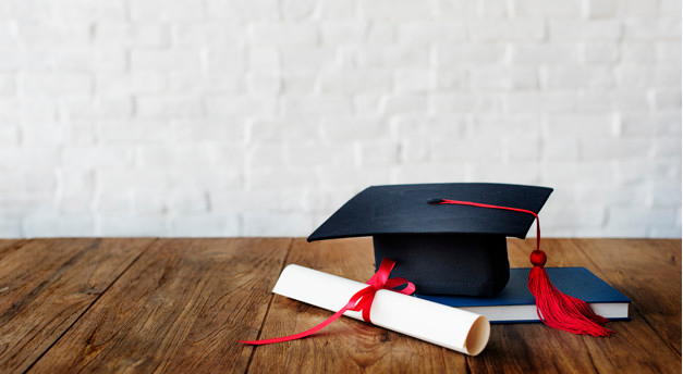 em uma mesa de madeira está um anuário, um chapéu de formando na cor preta e um diploma amarrado com laço vermelho