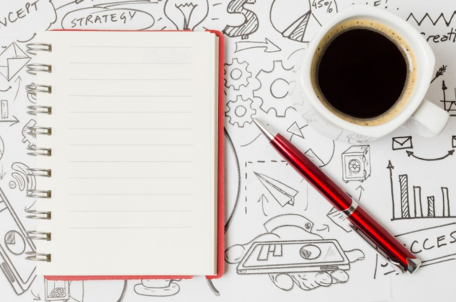 imagem de cima de uma mesa com um caderno, uma caneta e uma xácara de café