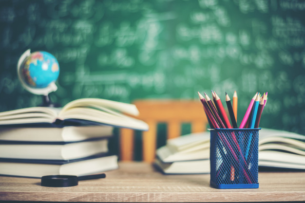 mesa de sala de aula com livros, canetas, um globo em miniatura e a lousa ao fundo