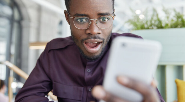 homem de camisa e óculos sentado com olhar de espanto e alegria enquanto encara seu celular