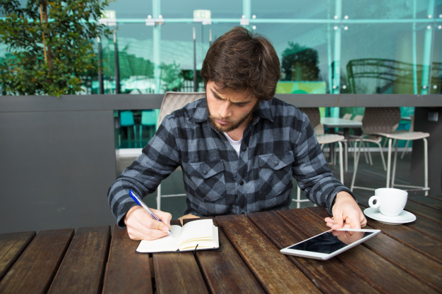 homem de camisa xadrez sentado a mesa de um café segurando um tablet com a mão esquerda e fazendo anotações com a mão direita em um bloquinho