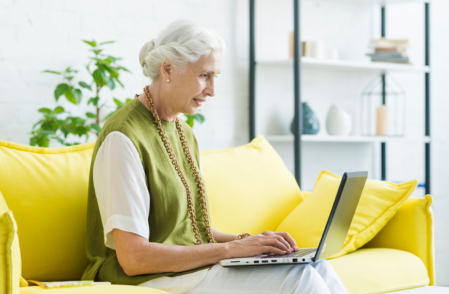 mulher idosa de colete verde sentada em um sofá amarelo usando um laptop