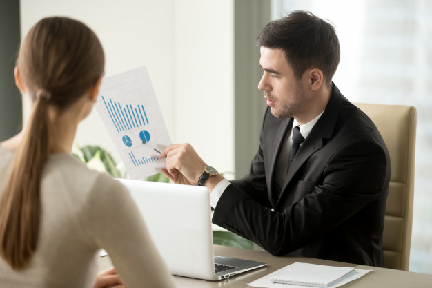 imagem de homem de terno sentado a uma mesa com uma mulher mostrando um gráfico em uma folha de papel
