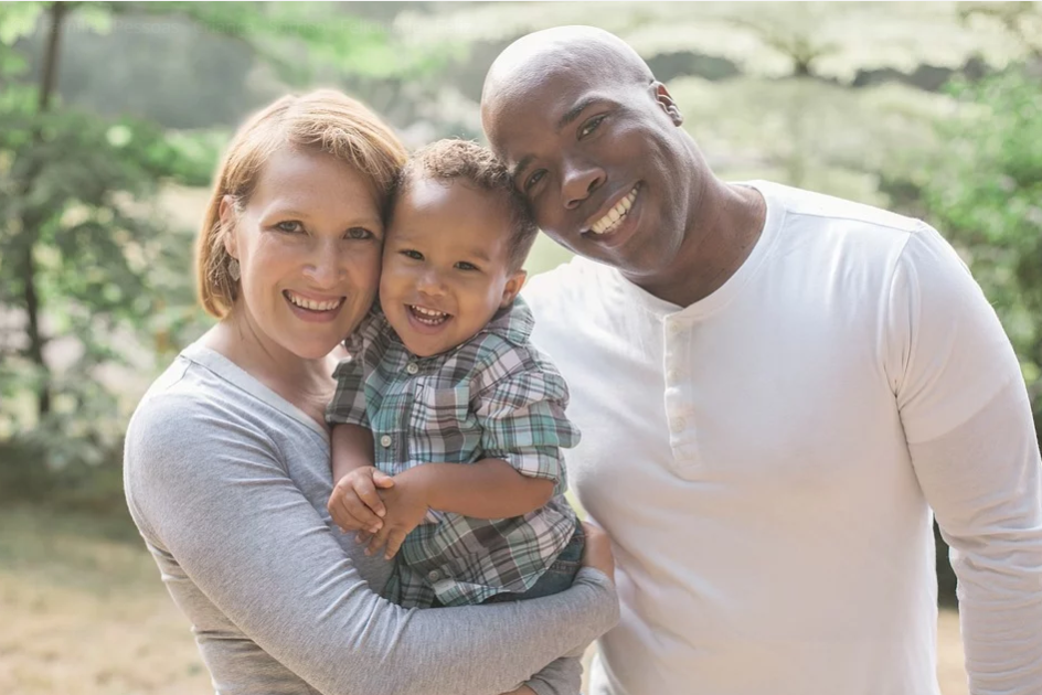 Casal feliz e sorrindo. No colo há um bebê filho deles. A mulher é branca e o homem é negro. 