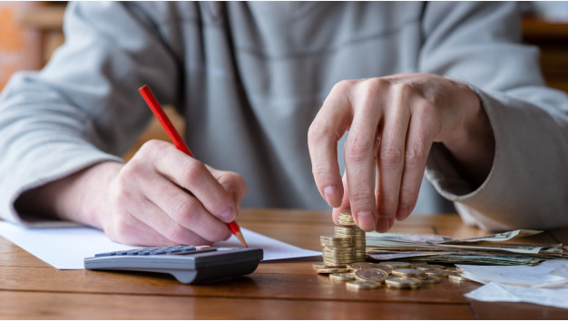 imagem ampliada de um homem contando moedas em uma pilha e fazendo anotações em uma folha ao lado de uma calculadora