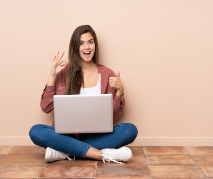 garota sentada no chão com seu computador no colo fazendo sinais positivos com a mão