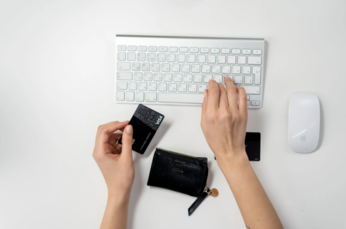 imagem da mão de uma pessoa segurando cartão e mexendo no teclado que está em cima de uma mesa e entre as mãos tem uma carteira 