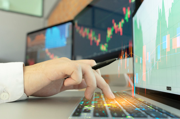 imagem ampliada de mão de um homem usando um teclado em frente a 3 telas com gráficos com dados sobre valor de ações