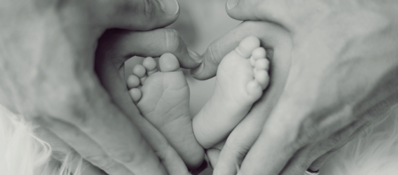 mãos do pai e mãos da mãe ao redor dos pés de um bebê em formato de um coração