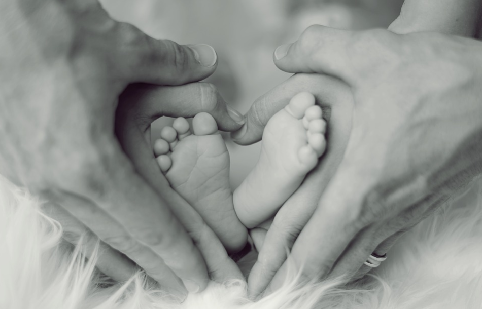 mãos do pai e mãos da mãe ao redor dos pés de um bebê em formato de um coração