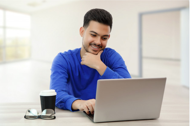 homem de camisa azul sorrindo enquanto leva a mão ao queixo sentando a uma mesa com um copo de café e um laptop