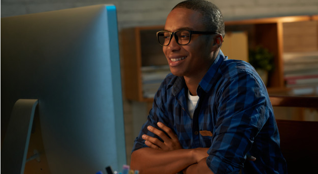 homem de camiseta xadrez azul e preta usando óculos sentado a uma mesa observando um monitor