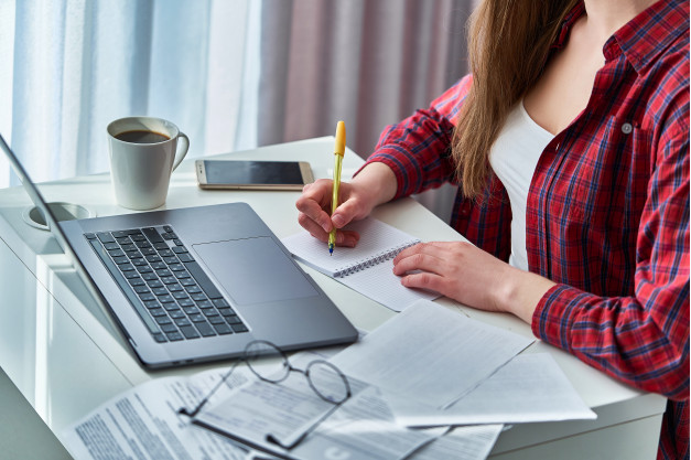 imagem ampliada de uma mulher usando camisa xadrez vermelha e preta fazendo anotações em um bloco de notas enquanto está sentada a uma mesa com folhas um par de óculos e um laptop