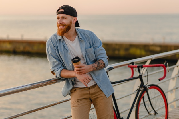 homem com bicicleta e copo de café sorri apoiado na grade de uma ponte acima de um rio