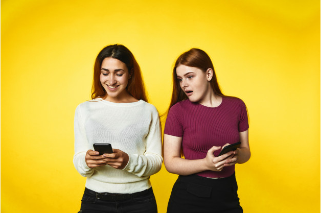 de costas para um fundo amarelo duas mulheres uma olhando o celular e a outra olhando para o celular da amiga com olhar de ciúmes