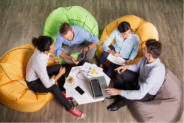 grupo de quatro amigos sentados em puffs com uma mesa de centro e um laptop vistos de cima